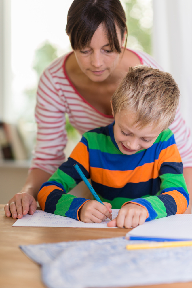 Concerned mother looking over the shoulder of a young boy as he sits at a table writing or drawing on a sheet of paper